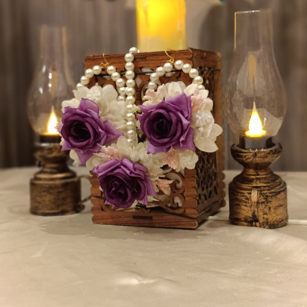 A set of floral earrings and a bindi displayed on a decorative box with a candle lit in the background. The earrings are adorned with artificial flowers in purple and white colors.