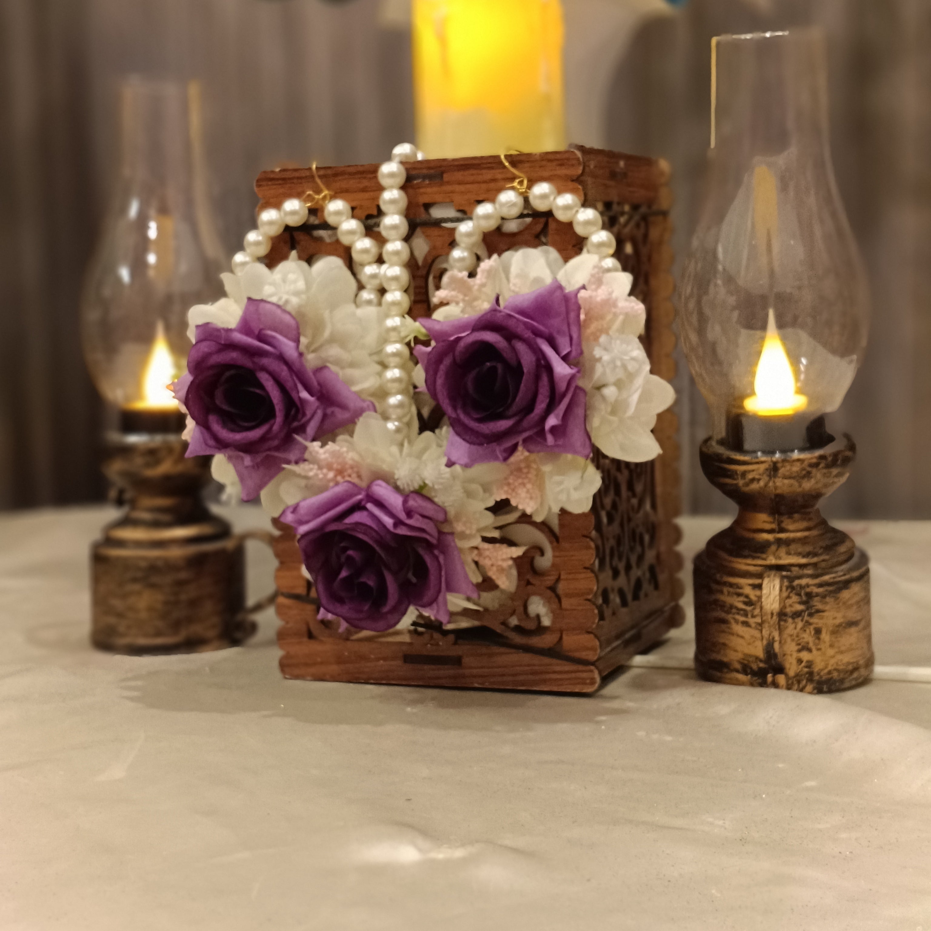 A set of floral earrings and a bindi displayed on a decorative box with a candle lit in the background. The earrings are adorned with artificial flowers in purple and white colors.