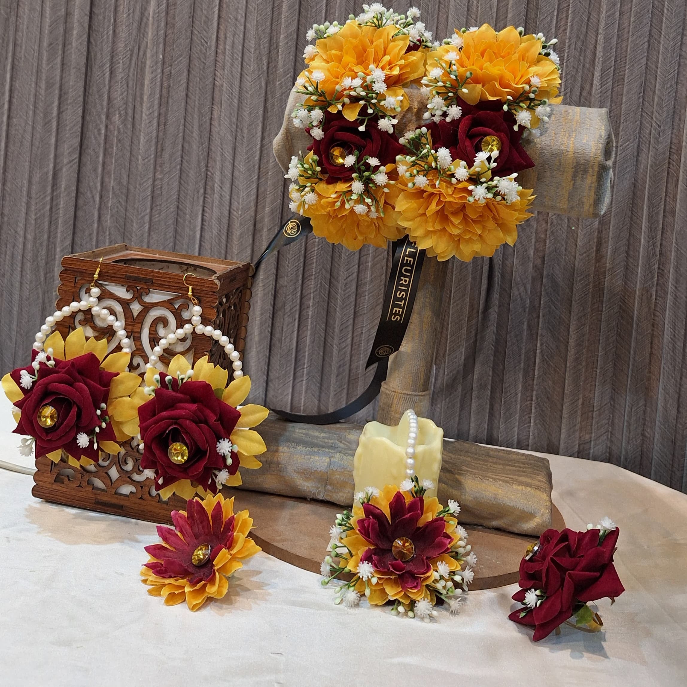 Decorative floral arrangement with maroon and yellow flowers on a wooden stand against a textured wall.